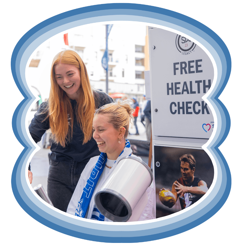 Two women laughing at a Shane Warne Legacy free health check station at a sporting event